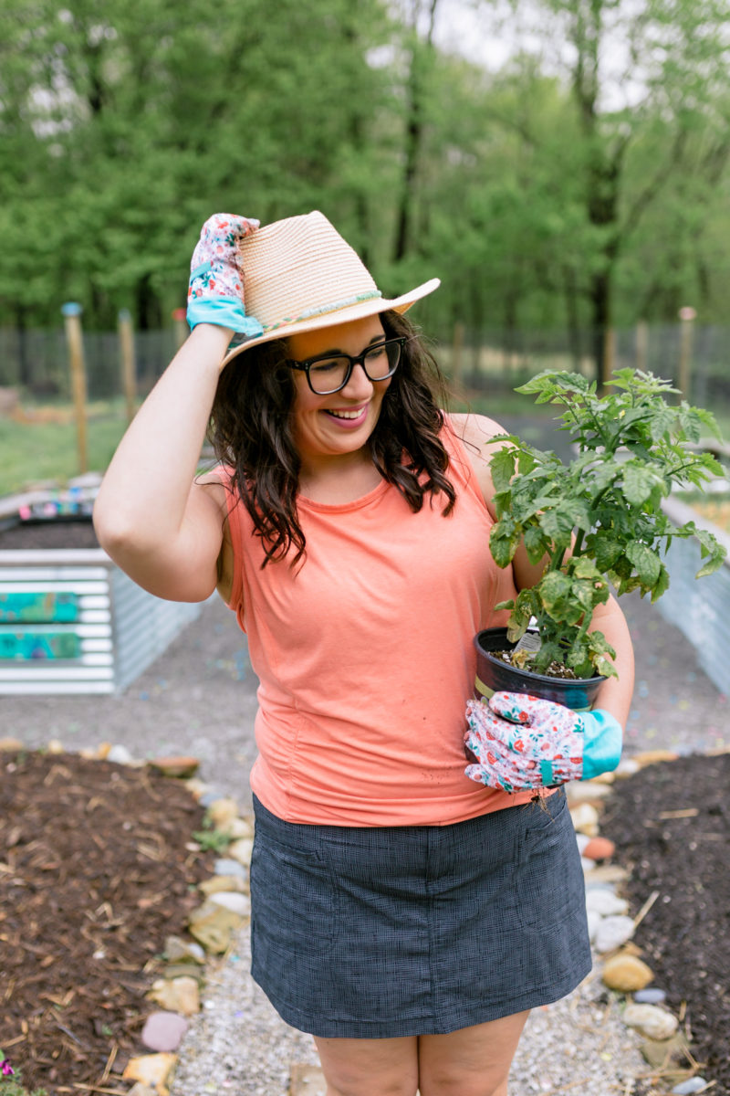 A brunette woman stands in a garden smiling and holding a tomato plant.