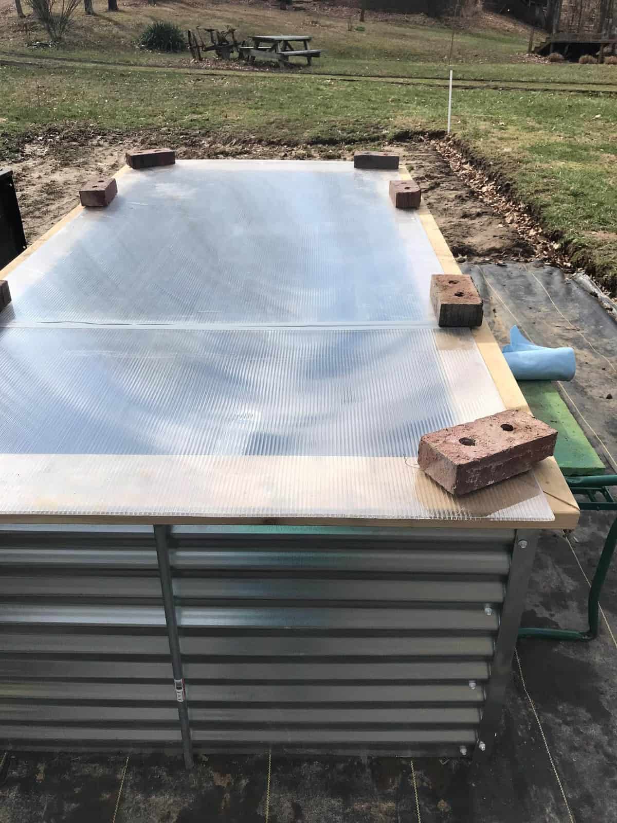 Side shot of a Galvanized Steel Raised Garden Bed with a greenhouse panel laid over top, held down with bricks