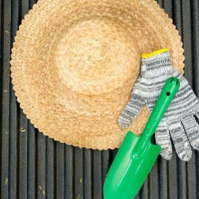 A green trowel and a striped garden glove sits on top of a straw gardening hat