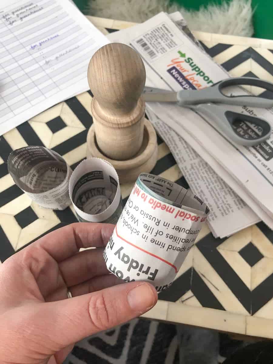 A hand holds a small pot meant for seedlings, made out of newspaper. Newspaper, scissors, and a pot maker are in the background.