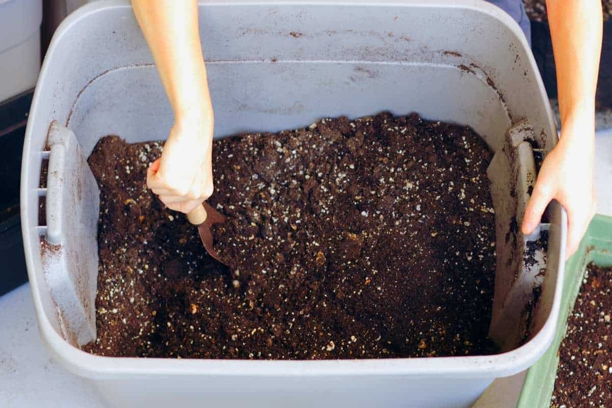 A hand uses a trowel to stir components for soil for indoor plants in a large tub.