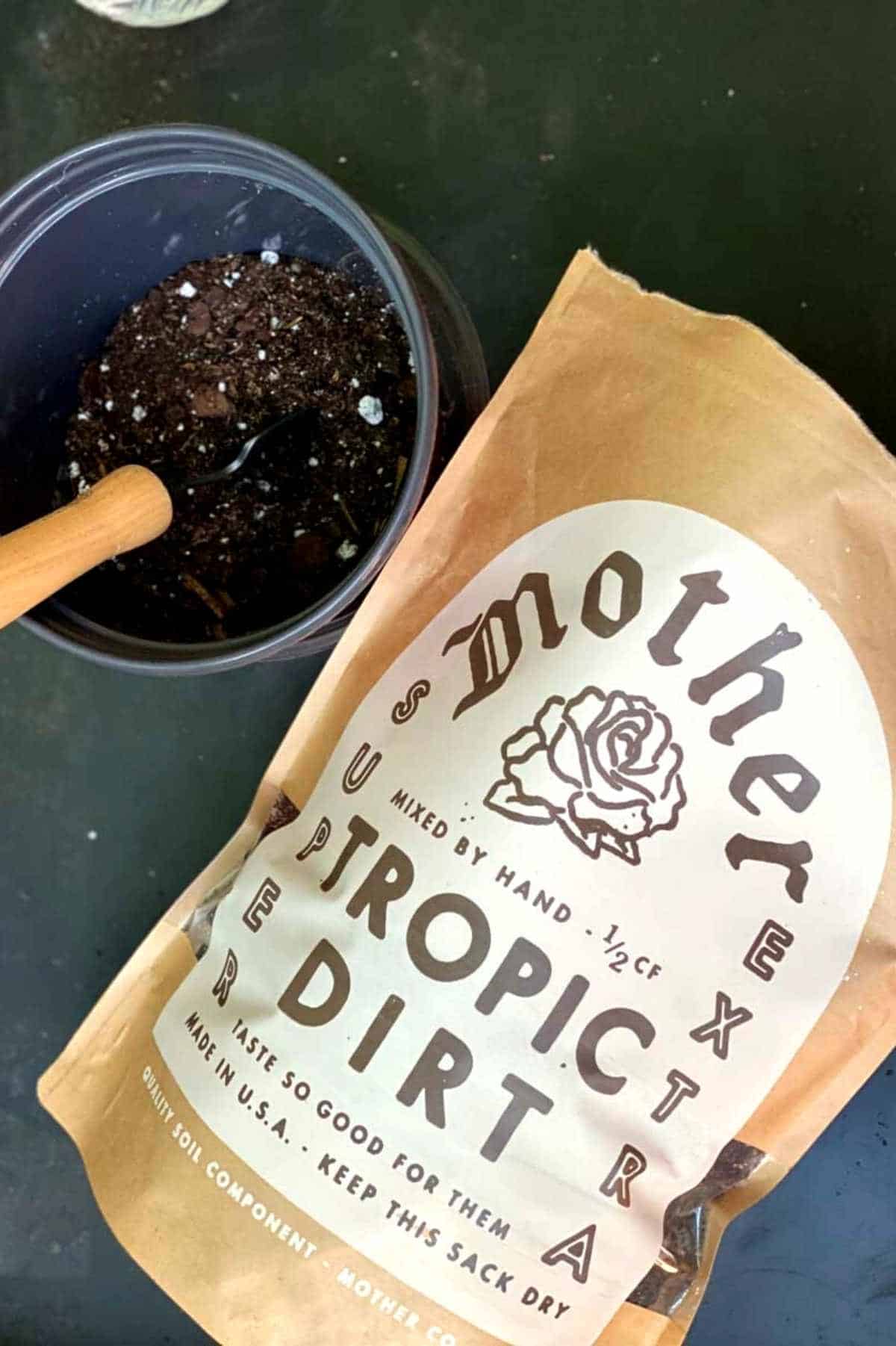 A bag of Mother Co. tropic dirt soil for indoor plants next to a container and trowel